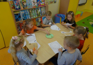 Sześcioro dzieci siedzi przy stoliku, w ręku trzymają nożyczki i wycinają tarcze zegarów z papieru.